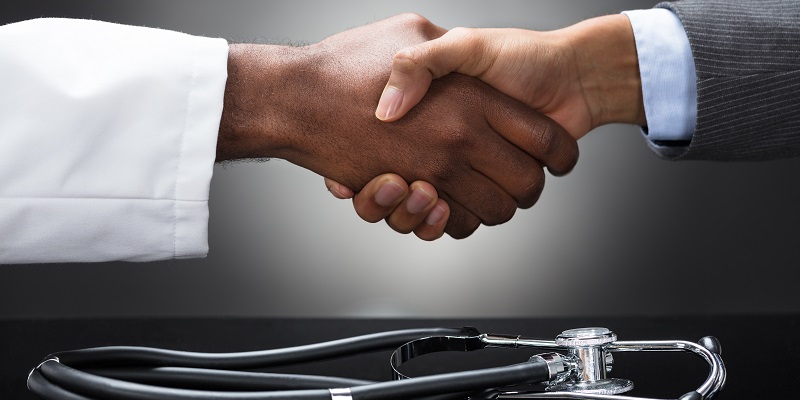 Two hands shaking across a black table with a stethoscope sitting on it.