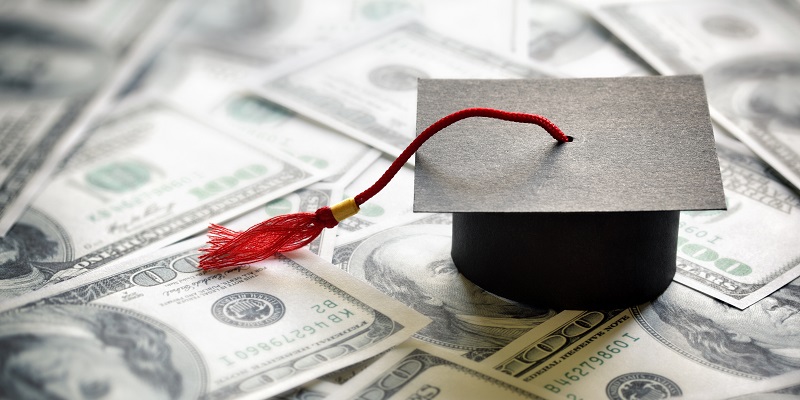 A small graduation cap sits on top of a pile of $100 US bills